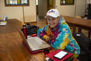 A woman in a tie-dye hoodie and baseball cap smiles at the camera while working on her laptop at a long wooden table.