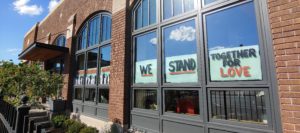 Front face of the Green Garage, a historic building made of brown brick with large square and arch-shaped windows. The square windows show painted signs that say “We Stand Together For Love.”