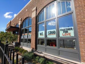 Front face of the Green Garage, a historic building made of brown brick with large square and arch-shaped windows. The square windows show painted signs that say “We Stand Together For Love.”