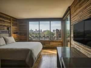 A cozy room with a large bed, concrete countertops, repurposed wood walls, a wall-mounted TV, and large windows looking out onto the Detroit skyline.