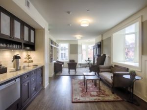 A handsomely furnished room with historic wood floors, modern appliances, large windows, and historic furniture: the parlor of the El Moore. Kitchen counter, couch and side tables are in the foreground. In the background is a couch, 3 chairs and a seating area around a round table.