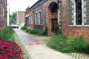 A linear path made of concrete, brick, and permeable pavers bordered by lush gardens alongside the Green Garage, a historic brick building with numerous windows. Red flowers and tall grasses are shown.
