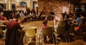 25 diverse people sitting in a large drum circle in the center of the Green Garage, playing various-sized drums together.