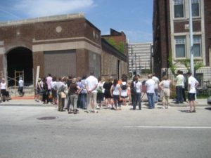 Large group of people gathered on the street, just next to the Green Garage, listening to a community member describing the construction process. Some windows are bricked up; one window opening is open to the elements.