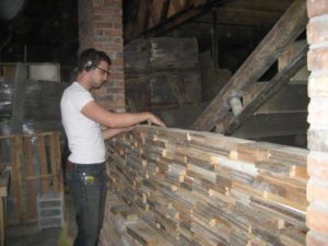 A man places pieces of discarded wood in place in the formation of a wood wall next to the front staircase at the Green Garage, surrounded by various construction materials.