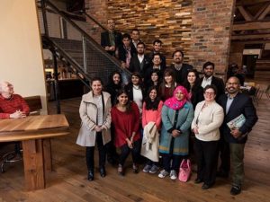 20 people visiting the Green Garage pose in front of a 2-story wood wall, standing in tiers along a staircase, while a man at a wood reception desk smiles up at them.