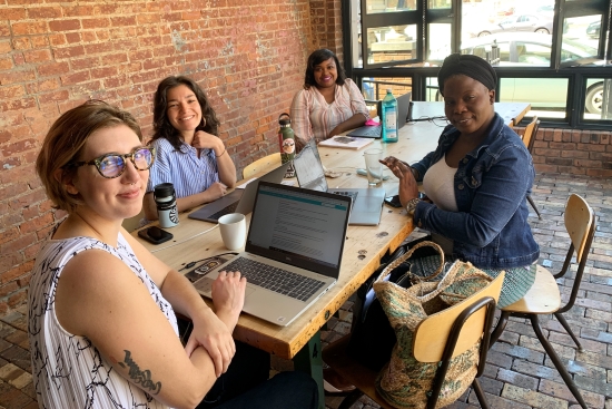 4 diverse women smiling and sitting at a table in a brick indoor/outdoor room, holding a meeting. Windows open in the background.