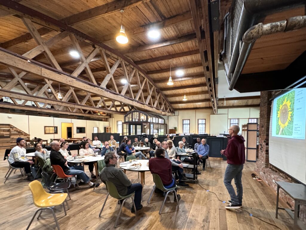A man giving a slide presentation to 25 people sitting at 6 circular tables in a large open room with wood floors. Above the crowd is a high wood ceiling, historic bow trusses, pendant lights, and Solatubes, which let natural light into the building.