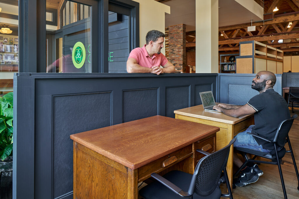 2 men talking at a 2-person office. One man is sitting at his computer, and the other is standing on the outside of chest-high space divider. A spacious, open interior is seen behind them.