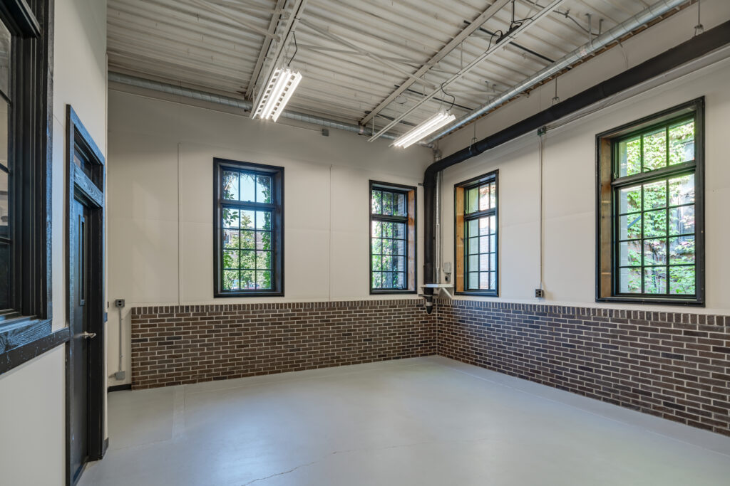 Closed-door workshop space with 4 large windows on 2 walls, overhead lighting, concrete floor, and bricks lining the lower walls under the windows.