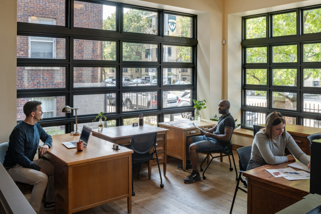 3 people work in a corner office lined with exterior windows on 2 sides. Each person works at their own desk, and a 4th desk sits empty.