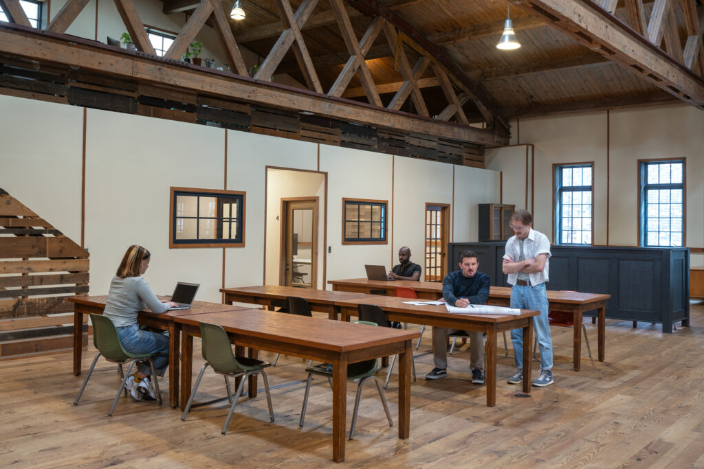 4 people working independently in an open area at 3 long tables with seating for 12. Conference rooms and exterior windows are seen in the background.