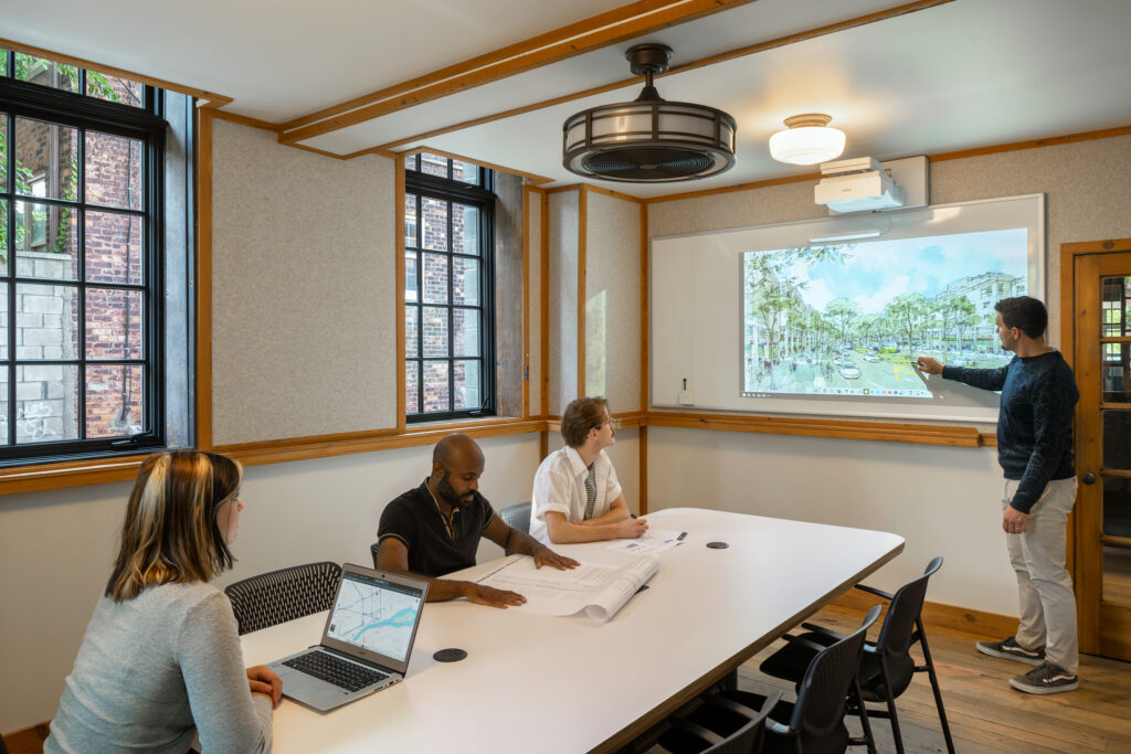 Meeting in large conference room. 4 community members working together, 3 seated at large white table and 1 standing at large whiteboard with architectural drawing displayed. 2 windows in background.