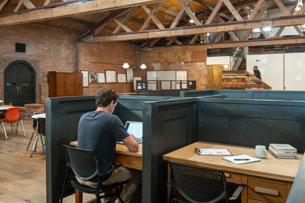 A man sits at an individual desk in his one-person office, separated by an adjacent office by 3-ft high black dividing panels. A spacious, open, wood and brick interior is seen around him.
