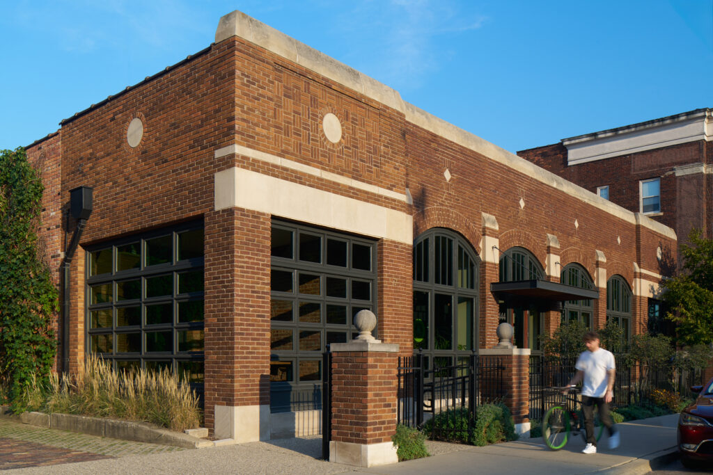 Exterior angled photo of Green Garage’s historic brick facade on a cloudless day, showing brick posts and iron fence, grasses, shrubs, climbing plants, a tree, and a man walking his bike past the building.