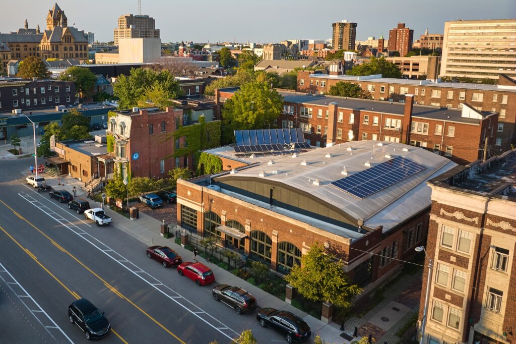 The Green Garage in its neighborhood context from above, showing the white barrel vault roof and solar panels. Other buildings shown include the Bronx Bar, apartment buildings, and Old Main, a historic building at Wayne State University.