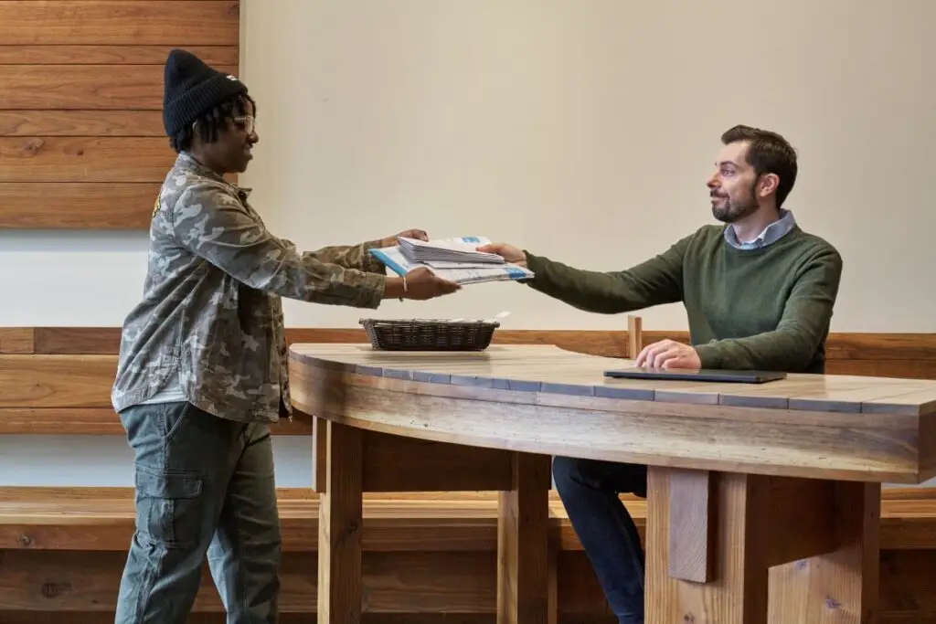 A man seated at a custom-built wood desk hands a stack of mail to a woman standing on the other side of the desk.