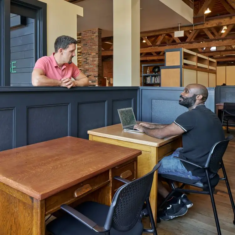 2 men talking at a 2-person office. One man is sitting at his computer, and the other is standing on the outside of chest-high space divider. A spacious, open interior is seen behind them.