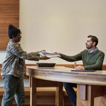 A man seated at a custom-built wood desk hands a stack of mail to a woman standing on the other side of the desk.