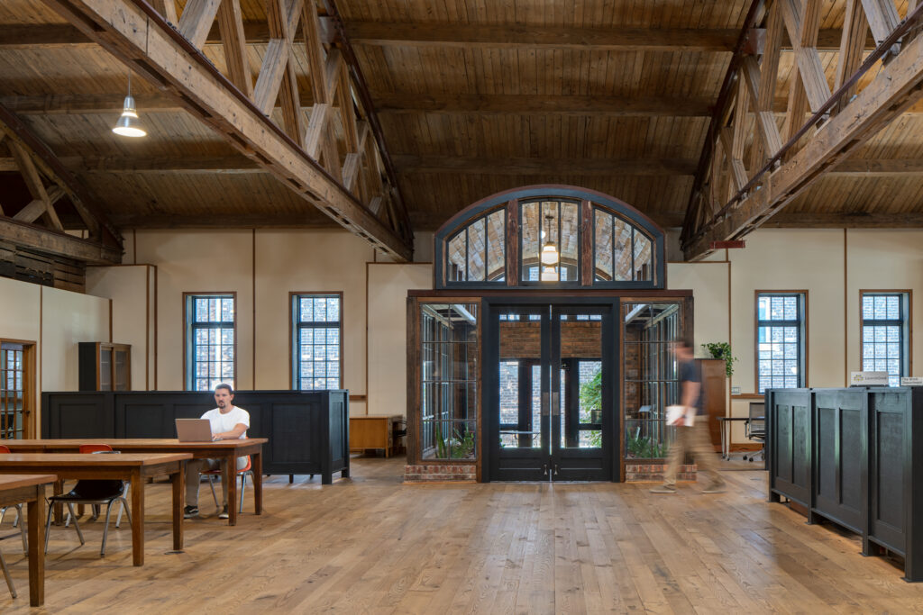 A photo of a large open space with wood floors, wood tables, and a curved wood ceiling, with large trusses under the ceiling. A man sits at one of the tables with his laptop open.